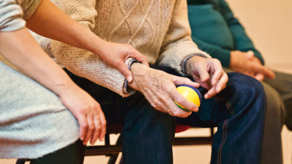 An elderly man is holding a stress ball, and a woman is holding his hand